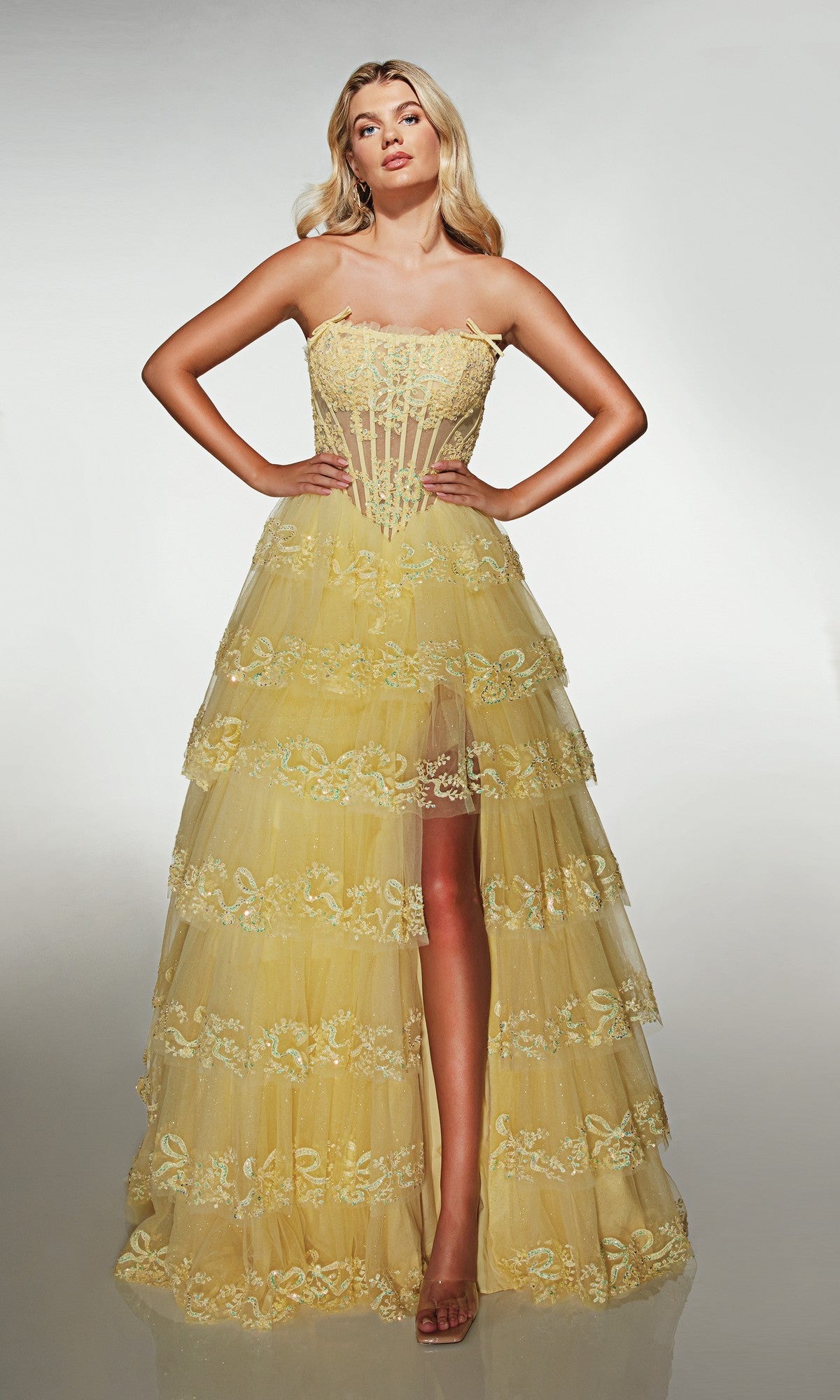 A woman models the Alyce 62191 long prom dress, featuring a strapless corset bodice and tiered yellow tulle A-line skirt with lace accents, standing hands-on-hips against a plain background.